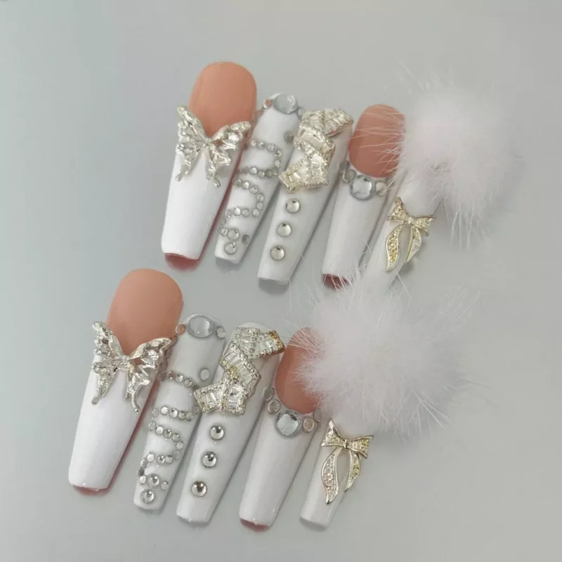 White and nude bridal nails with rhinestone bows, butterflies, and fluffy pom-poms