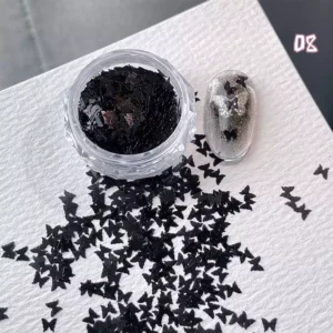 Black butterfly-shaped nail glitter spilled beside swatch tip on white paper