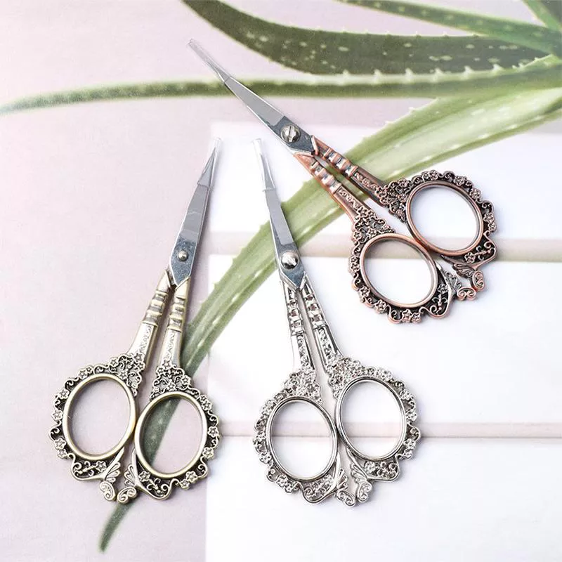 Three ornate vintage nail scissors in silver, gold, and rose gold finishes on floral background